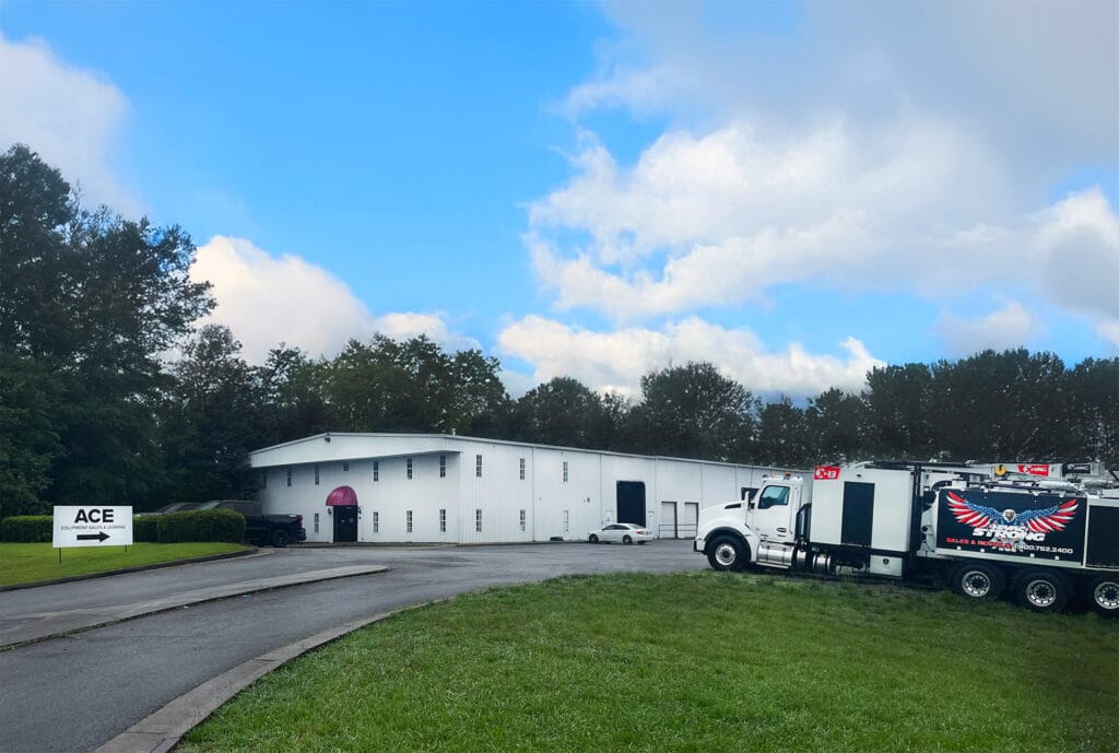 White industrial building and treeline under blue sky; X-Vac hydro-excavation truck parked right, ACE sign