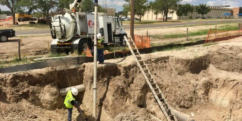 Two workers in a deep sandy excavation using an X‑Vac hydroexcavation vacuum hose and ladder beside the X‑Vac truck.