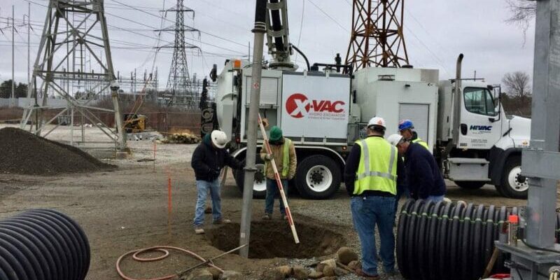 Workers in high-vis vests using X-Vac hydro excavation truck and vacuum boom to dig a utility hole at a power substation.