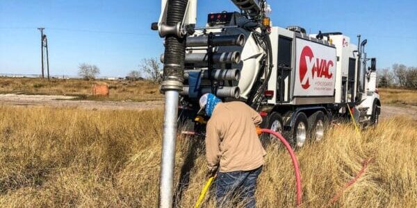 Worker handling hoses beside an X‑VAC hydroexcavation truck with boom and vacuum hose in a dry field